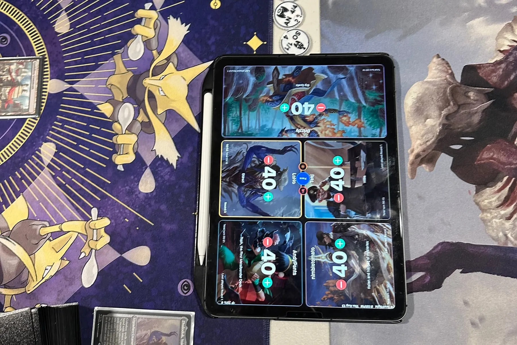 A Commander table mid-game with the TurnZero HQ life tracker running on a tablet, five seats at 40 life, a row of status tokens beside it