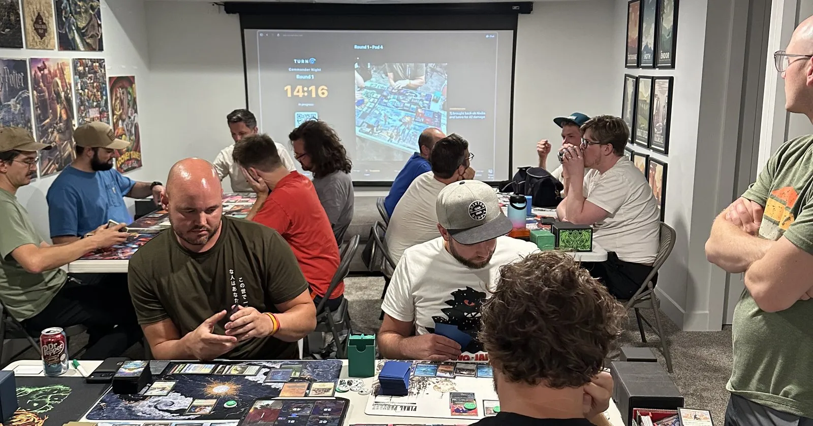 A dozen players at three tables mid-game, boards covered in cards, with TurnZero's Commander Night round clock projected on the wall behind them reading Round 1 with fourteen minutes left.