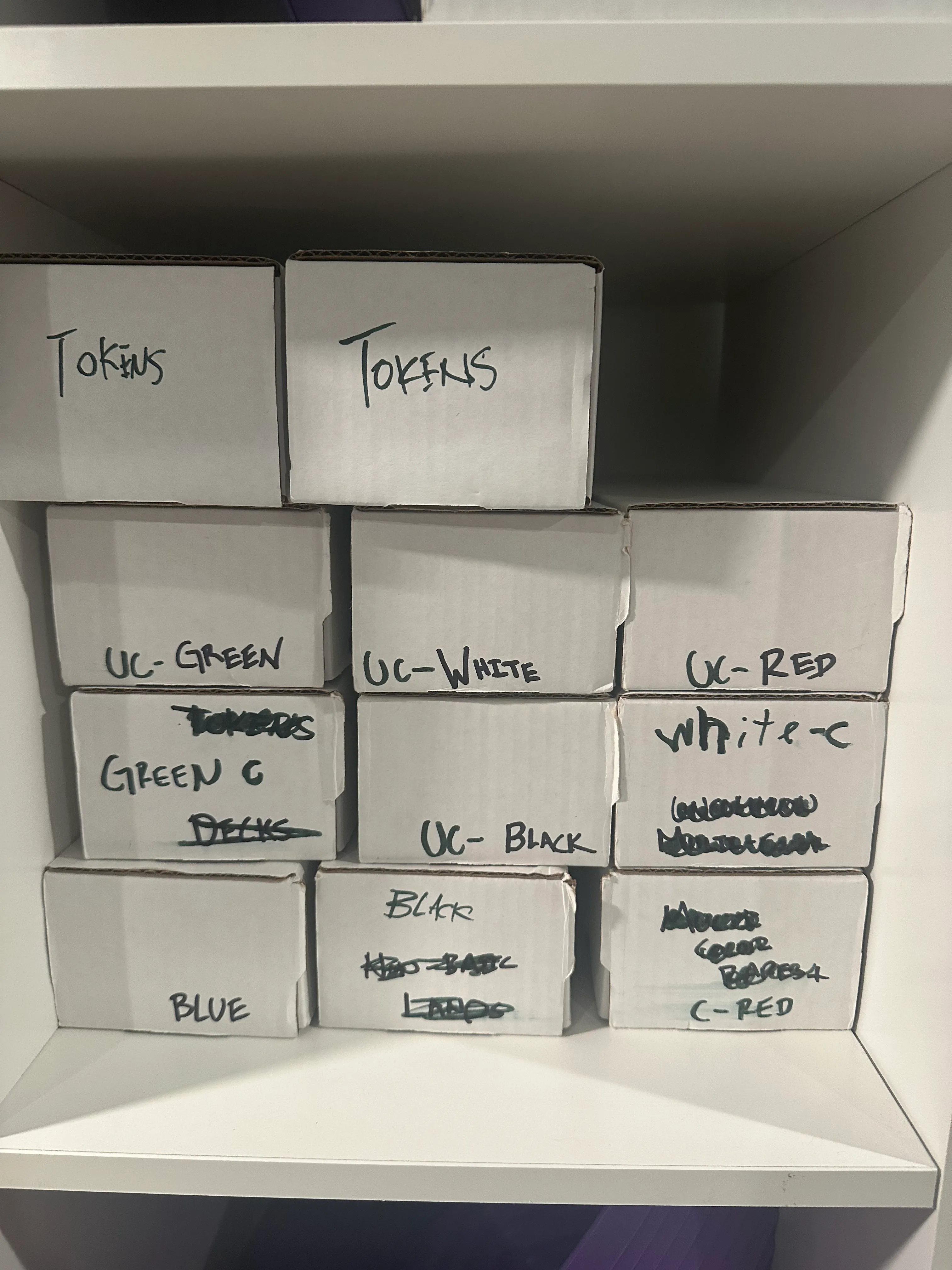 A shelf of labeled cardboard storage boxes for a card collection, marked by color (UC-Red, UC-White, UC-Black, UC-Green) and category (Tokens, Blue, Lands).