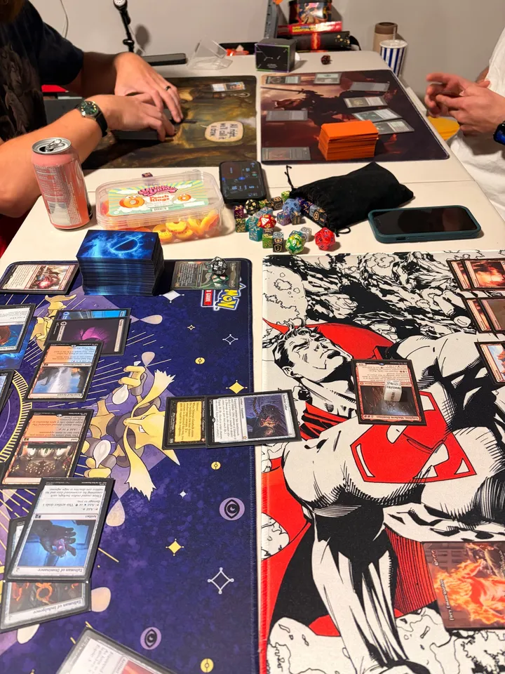 A casual Commander game at a kitchen table, phone tracker in the middle, next to a playmat that reads 'All right, we'll call it a draw'