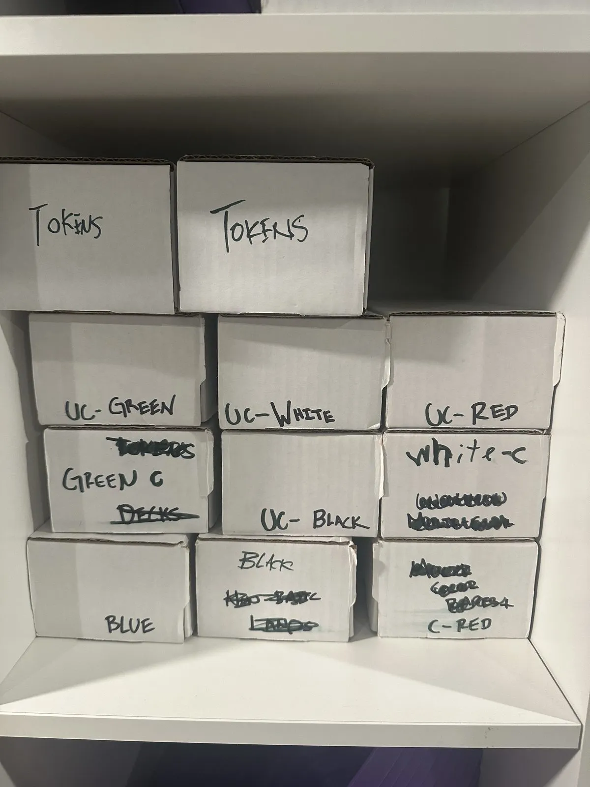 Eleven bulk boxes on a shelf, hand-labelled in marker by color: tokens, uncommon green, uncommon white, uncommon red, blue, black and common red, several of the labels crossed out and rewritten.