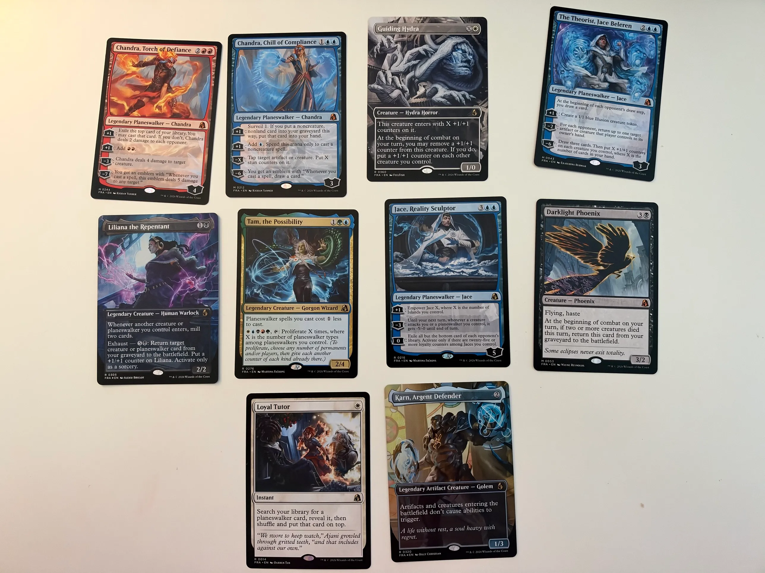 Ten Reality Fracture cards laid out on a table: The Theorist, Jace Beleren, Darklight Phoenix, Chandra, Torch of Defiance, Chandra, Chill of Compliance, Guiding Hydra, Liliana the Repentant, Tam, the Possibility, Jace, Reality Sculptor, Loyal Tutor and Karn, Argent Defender.